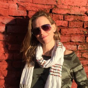 Person wearing sunglasses and a scarf stands against a red brick wall in daylight.