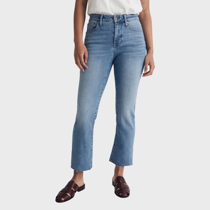Person wearing light blue jeans, standing casually in brown sandals, with a white shirt in a plain environment.