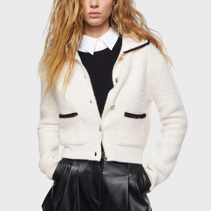 Woman wears a white cardigan over a black sweater and a white collared shirt, with hands in black leather pants pockets, against a plain background.