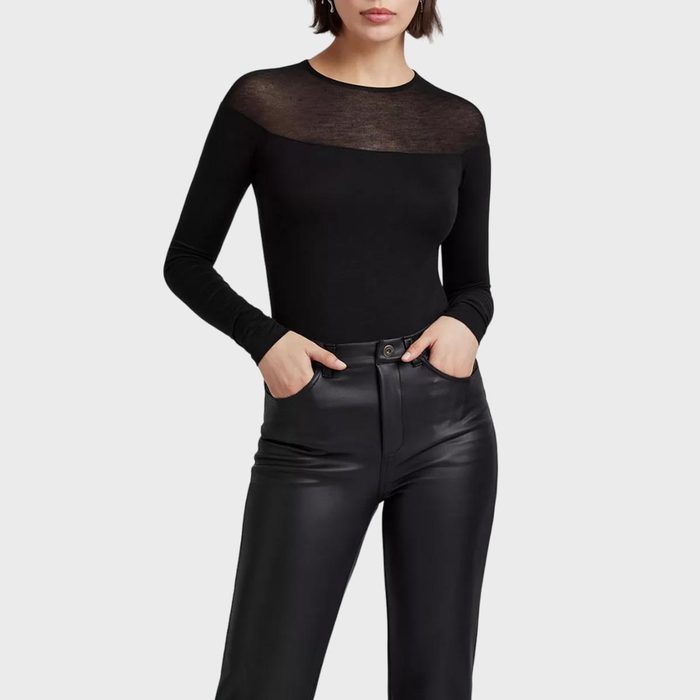 Person wears a black long-sleeved top with sheer neckline, hands in front pants pockets against a plain white background.