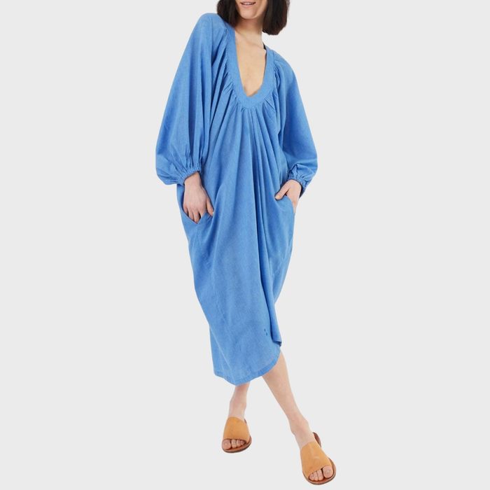 A person wears a loose, blue dress with long sleeves and brown sandals, standing against a plain white background.