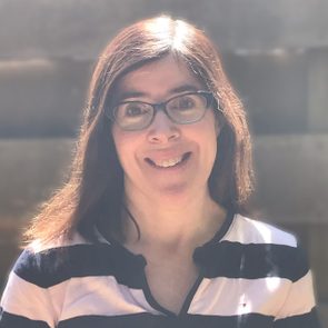 A person smiles, wearing glasses and a striped shirt, with sunlight illuminating their hair against a blurred, neutral background.