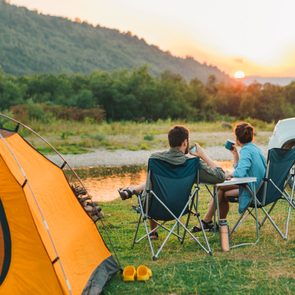Two people sit in camp chairs, drinking and enjoying the sunset next to an orange tent and white SUV near a river and forested hills.