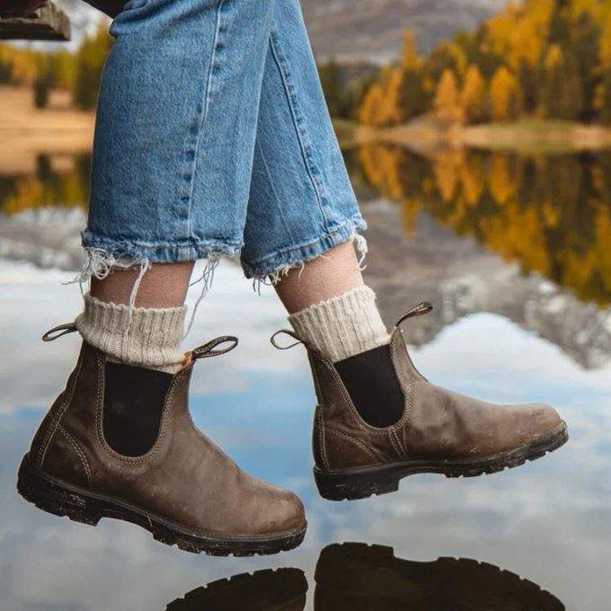 Rigid, worn boots rest above reflective water, framed by autumnal forest and mountains.