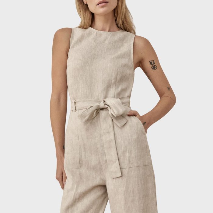 A person wearing a sleeveless beige jumpsuit with a waist tie stands against a plain background, showing arm tattoos.