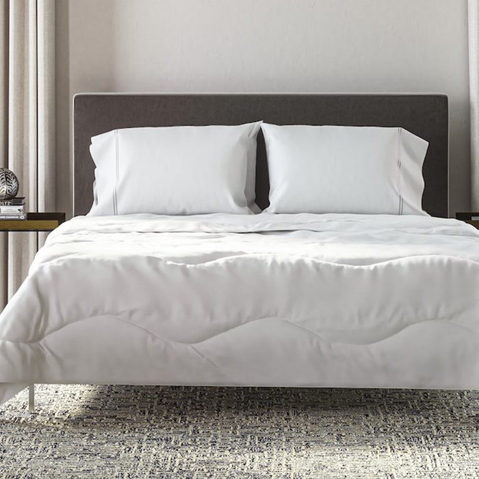 Bed is neatly made with white bedding and pillows, set in a modern room with light curtains and side tables holding books and decor.