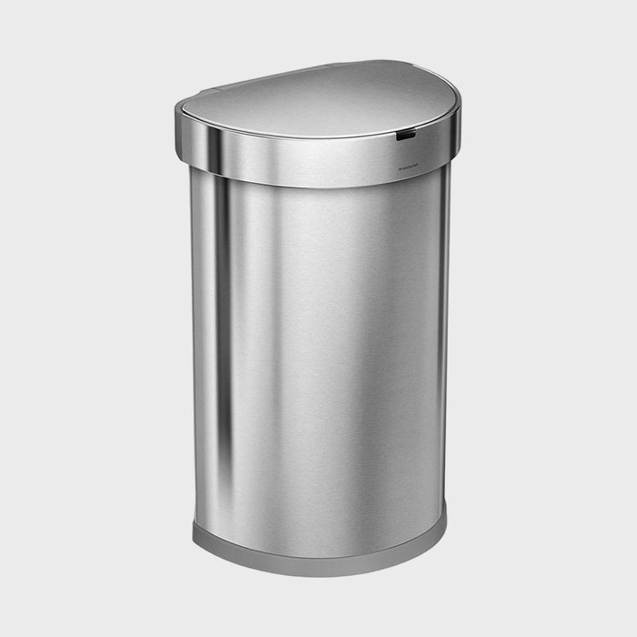 The stainless steel trash can stands upright, featuring a semi-round lid, in a plain, light-colored setting.