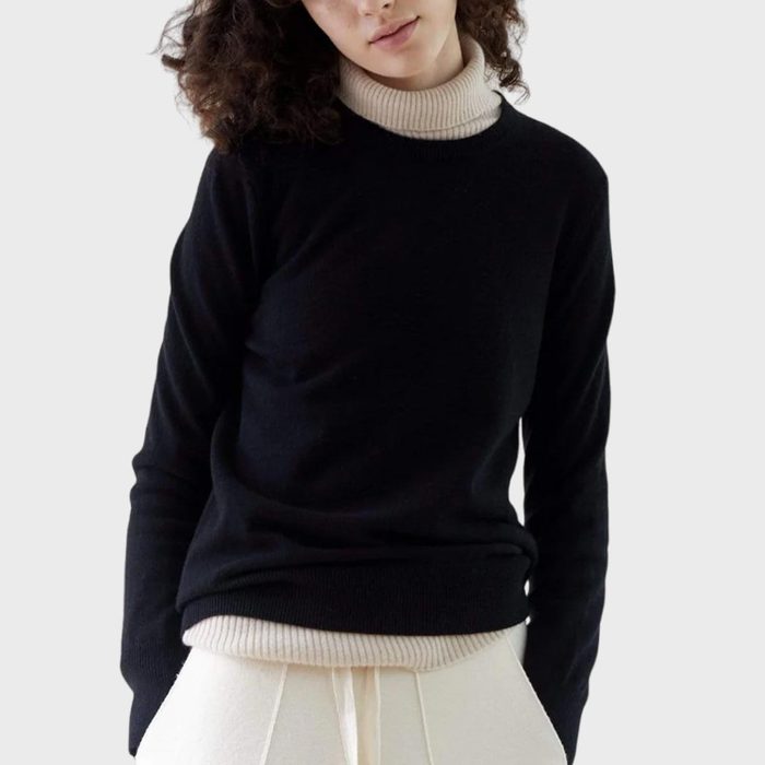 A person wears a black sweater over a beige turtleneck, standing against a plain grey background.