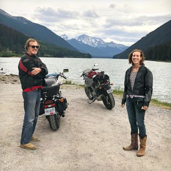 Two people stand by parked motorcycles, smiling, near a lake surrounded by forested mountains. License plates read "Y81477" and "3112."