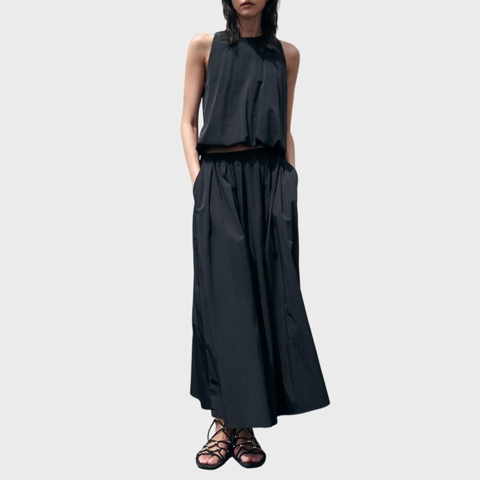 Person wearing a sleeveless, loose-fitting black outfit stands with hands in pockets, feet in strappy sandals, against a plain gray background.
