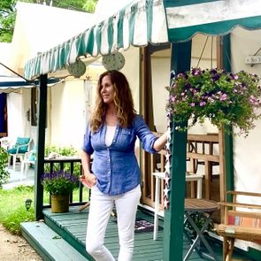 Woman stands smiling on porch, surrounded by potted plants, porch chairs, and a hanging basket of flowers. An American flag and 