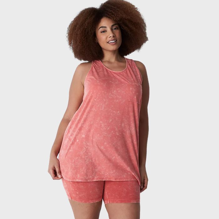 A person smiles, wearing a pink tank top and shorts, standing against a plain gray background.