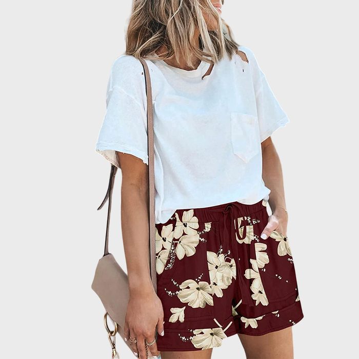 Woman wearing a white t-shirt and floral shorts holds a beige purse, standing against a plain background.