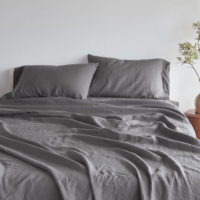 Gray bedspread lies wrinkled atop a neatly made bed, flanked by two pillows, with a potted plant on a nearby wooden nightstand.