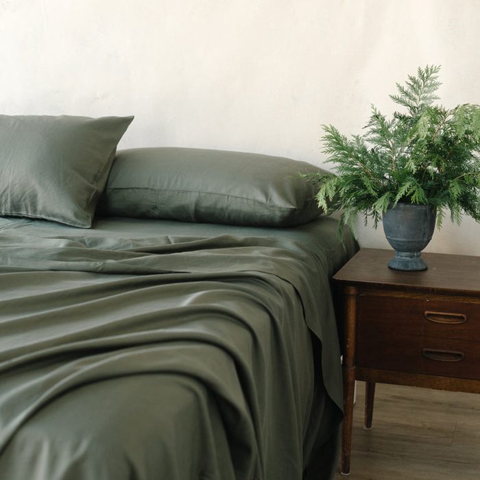Bed with green sheets and pillows, next to a wooden nightstand with a potted plant, set against a plain wall.