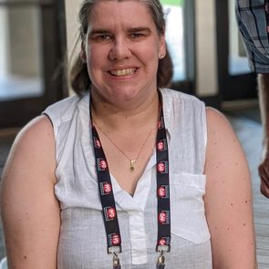 A person wearing a lanyard smiles, seated indoors near large windows. The name badge reads "Elizabeth Doughman, Managing Editor, Mitchell Media."