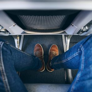 Feet wearing brown shoes rest under an airplane seat, surrounded by blue carpet and denim-clad legs.