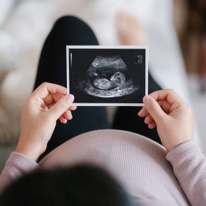 Hands hold ultrasound photo, viewed from above, over a pregnant belly in a cozy, softly lit setting.