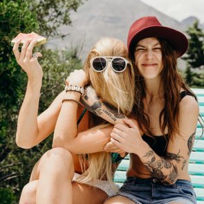 Two people sit smiling and embracing; one holds watermelon. Sunny outdoor setting with greenery, tattoos visible on arms, wearing casual summer clothing.