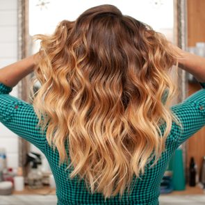 Wavy, ombre hair is being styled with both hands in a salon setting, surrounded by various hair products on shelves and countertop.