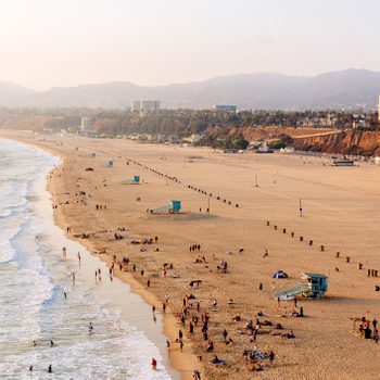 Beachgoers relax and play by the ocean, with sandy shores stretching towards distant mountains under a clear sky. Lifeguard towers and buildings visible.