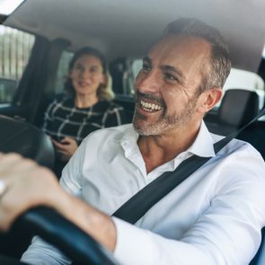 Man driving confidently, smiling while steering, with a woman sitting in the back seat, inside a car on a bright day.