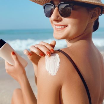 A woman applies sunscreen to her shoulder, smiling on a sunny beach with waves in the background.
