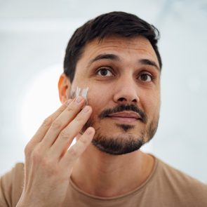 Man applies cream to his cheek in a softly lit bathroom, looking content.
