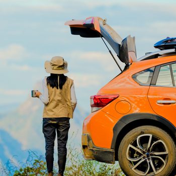Person stands drinking beside an orange car with open trunk, overlooking mountains, wearing a hat and vest.