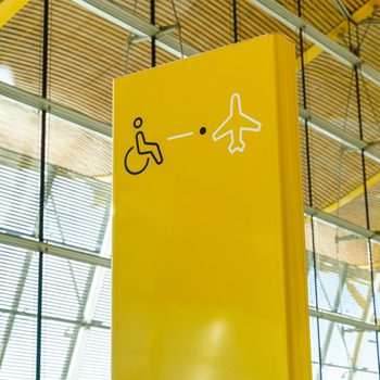 Yellow airport sign showing disability access directs toward plane, viewed in a terminal with glass and yellow structural elements. Gate J47 in background.