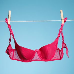 A bright pink bra hangs on a clothesline, secured with clothespins, set against a solid light blue background.