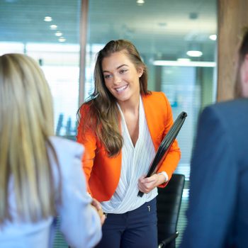 A woman shakes hands with two people, smiling, holding a folder in an office setting with glass walls and a conference table.