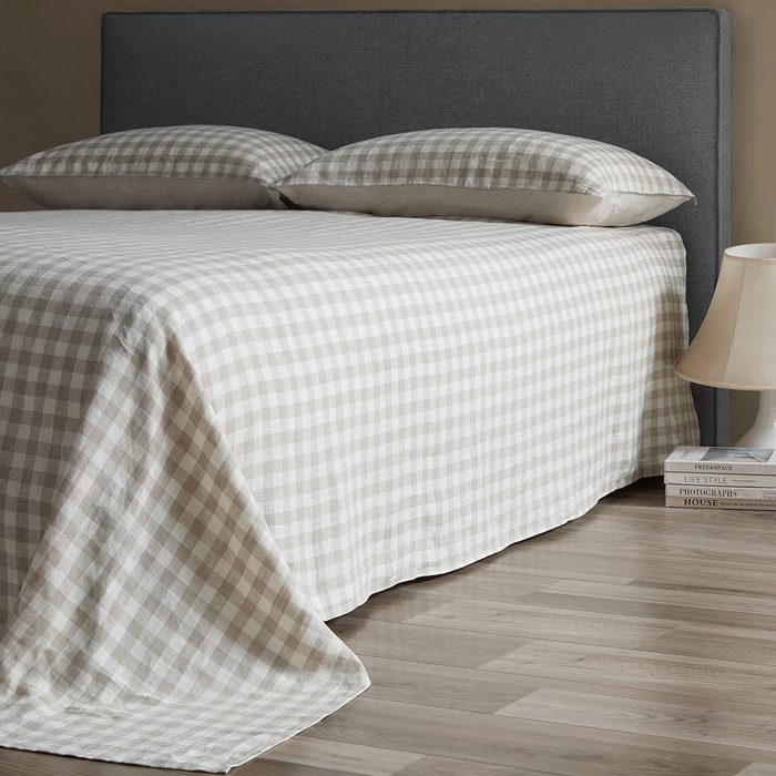 Bed covered in checkered sheets stands in a bedroom; two stacked books and a lamp sit on the floor beside it.