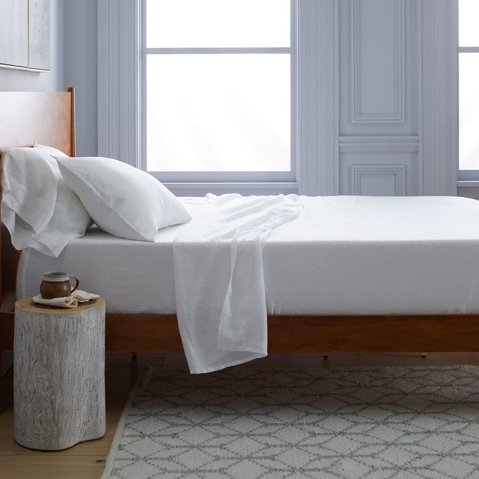 Bed with white sheets beside a rustic wooden table holding a mug, in a bright room with large windows and a patterned rug.