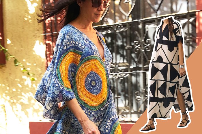 A woman walks, wearing a colorful, patterned dress and sunglasses, with a decorative metal fence and sunlight casting shadows behind her.