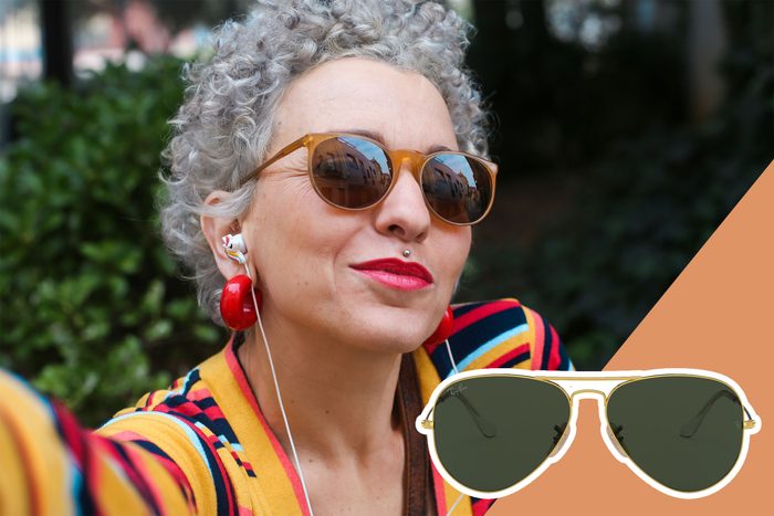 Person wearing sunglasses and colorful clothing smiles outdoors; mirrored sunglasses reflect buildings. Earphones and large red earrings are visible.