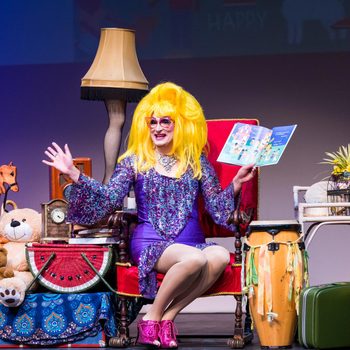 Person with bright wig reading a book, surrounded by toys and decor, seated on stage in a vibrant setting.