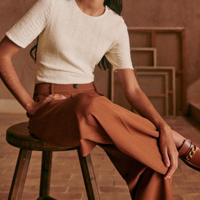 Woman sits on stool, wearing white top and brown pants, with art frames in the background.