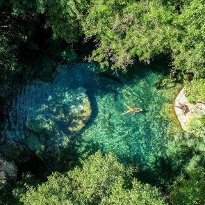 Person floats in a clear blue natural pool surrounded by lush, dense green foliage. Another person lounges on nearby rocks.