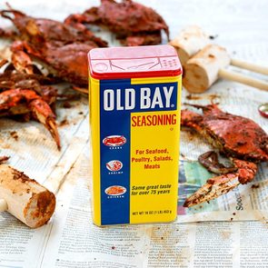 A container of Old Bay seasoning stands on newspaper with scattered crabs and wooden mallets nearby.