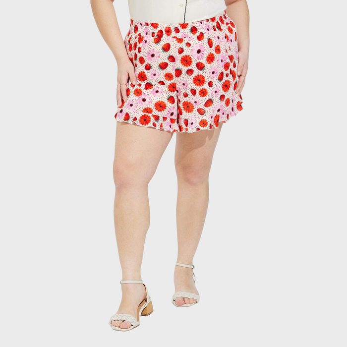 Woman wearing floral shorts stands, hands resting on hips, in a minimal white background. She wears white heeled sandals.