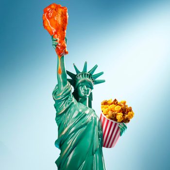 Statue of Liberty holds a giant dripping chicken leg and a striped bucket of popcorn against a light blue background.