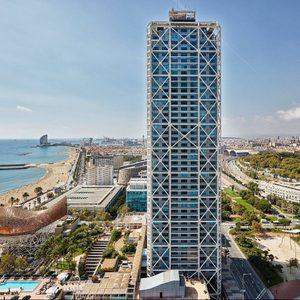 Skyscraper towers over beachside cityscape, featuring blue hues and crisscross design, surrounded by palm-lined streets and distant ocean views.