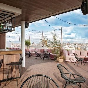 Rooftop patio with empty black chairs and pink seating, under string lights, surrounded by cityscape views and plants in daylight.