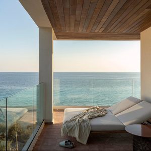 Lounge chair rests on a balcony overlooking a calm ocean, with a draped blanket under a wooden ceiling and glass railing.