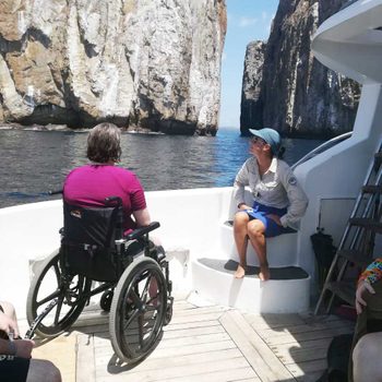 A person in a wheelchair faces majestic rock formations from a boat deck, accompanied by another person sitting on steps.