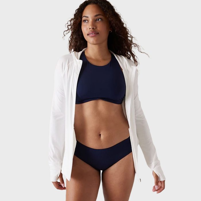Woman wearing a navy swimsuit and white jacket stands confidently against a plain, light gray background.