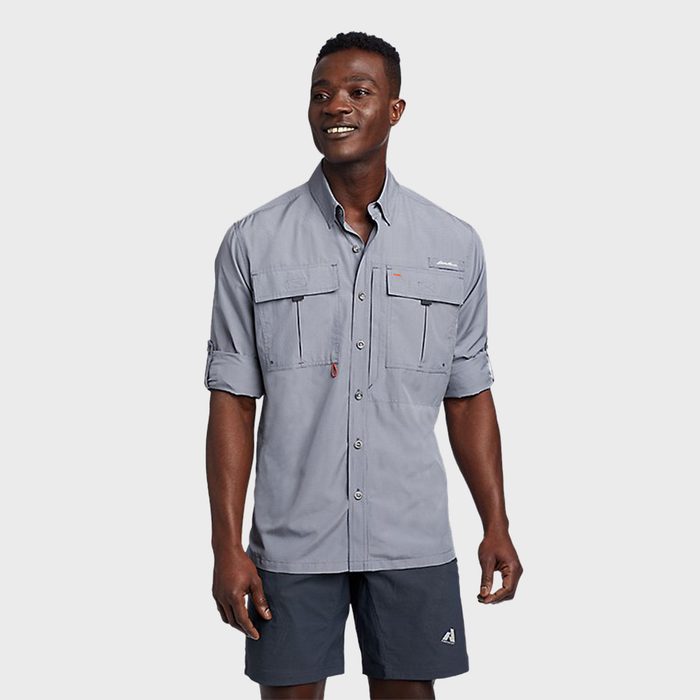 Man smiles, wearing a gray collared shirt with rolled sleeves and dark shorts, standing against a plain light gray background.