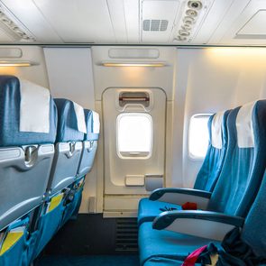 Airplane seats line the cabin, facing forward, with blue upholstery and white headrest covers, adjacent to windows and overhead compartments on both sides.