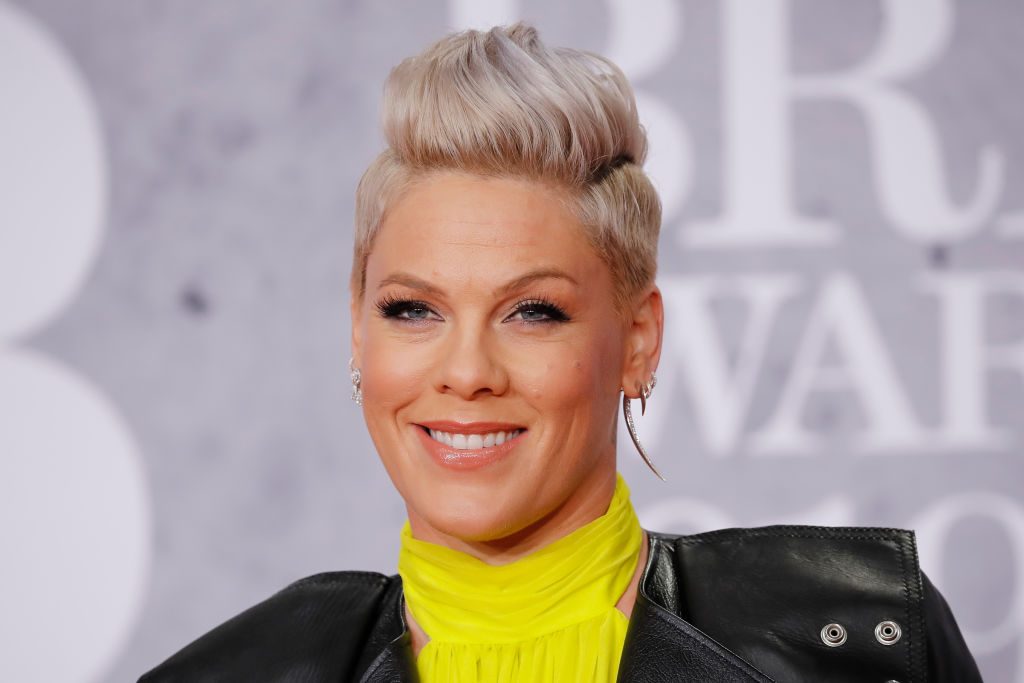 Person smiling, hair styled upwards, wearing bright yellow and black leather jacket, standing against a blurred background with partial text.
